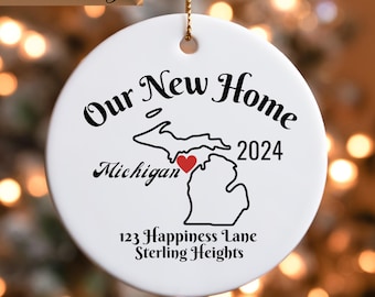 May include: White ceramic Christmas ornament with a black outline of the state of Michigan and the text "Our New Home 2024" with a red heart in the center of the state. The text "Michigan" is below the state outline. The text "123 Happiness Lane Sterling Heights" is below the state.
