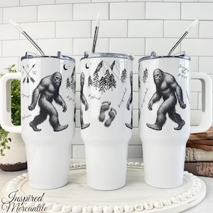 May include: Three white stainless steel tumblers with handles and straws, featuring black line art of Bigfoot, pine trees, mountains, footprints, and the text "Legend," "Believe," "Bigfoot," and "Simply the Best" printed on the exterior surfaces.