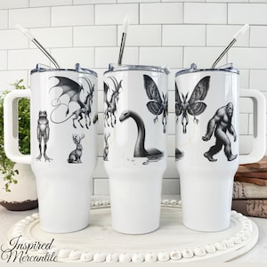 May include: White travel mugs with black and white illustrations of mythical creatures, including a frog, dragon, moth, and Bigfoot. Each mug has a white handle, a silver straw, and a white lid. The mugs are on a white tray.