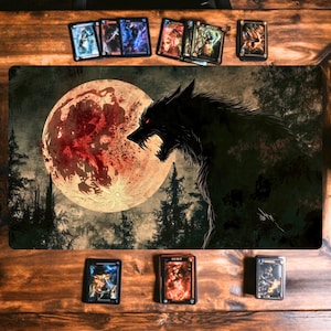 Puede incluir: Una alfombrilla de juego con una ilustración oscura de un lobo gruñendo contra una luna rojo sangre y un bosque. Varias cartas de juego de cartas coleccionables están dispuestas alrededor de la alfombrilla sobre una superficie de madera.