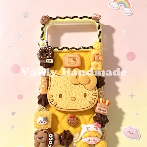Op de afbeelding: Een gele telefoonhoes versierd met 3D-harsbedels, waaronder een groot Hello Kitty-gezicht, een bruine beer met het label Coco, diverse koekjes, chocolaatjes en miniatuurvoedsel. De hoes heeft een doorgestikte textuur en donkerbruine biezen.