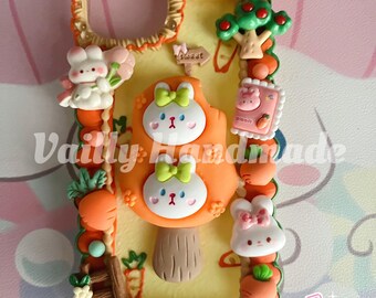 May include: A yellow phone case decorated with 3D resin charms including two white rabbits with green bows, orange carrots, a small tree, a wooden ladder, and a sign that says sweet. The case features a textured border and the text Vailly Handmade.