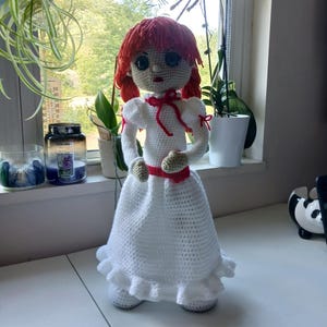 Może przedstawiać: Ręcznie robiona szydełkowa lalka z rudymi włosami z przędzy i białą sukienką. Lalka ma czerwoną wstążkę wokół talii i szyi. Ręce i dłonie lalki są beżowe. Lalka stoi na białej powierzchni.