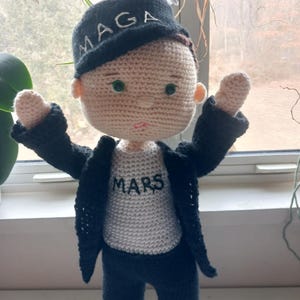 May include: A crocheted doll wearing a black jacket, black pants, and a white shirt with the word "MARS" on it. The doll is wearing a black hat with the word "MAGA" on it.
