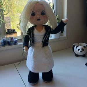 Könnte beinhalten: Handgemachte Häkelpuppe mit weißem Kleid, schwarzer Jacke und schwarzen Stiefeln. Die Puppe hat blondes Haar und einen strengen Gesichtsausdruck. Die Puppe steht auf einer weißen Oberfläche in der Nähe eines Fensters.