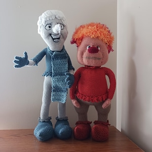 May include: Two handmade crochet dolls. One doll is tall with a white head, blue scarf, and blue gloves and boots. The other doll is shorter with orange hair, a red shirt, and red boots. Both dolls are standing on a wooden surface.