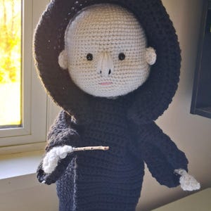 Puede incluir: Muñeca de ganchillo de un personaje con rostro pálido, túnica con capucha negra y varita. La cara de la muñeca tiene ojos negros, boca rosada y nariz blanca. La túnica y la capucha son negras.