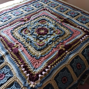 Puede incluir: Una manta cuadrada de ganchillo hecha a mano con un diseño floral y geométrico. La manta presenta un diseño central de diamante con un motivo floral, rodeado de bordes en tonos azules, rosas, beige y burdeos. Hecha de hilo suave.