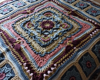 May include: A handmade, square crochet blanket with a floral and geometric design. The blanket has a central diamond pattern with a flower motif, framed by borders in blue, pink, beige, and burgundy. The blanket is made of soft yarn, perfect for a bed or sofa.