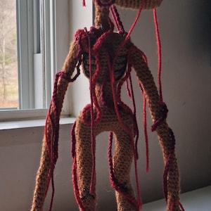 Op de afbeelding: Gehaakte Siren Head figuur in bruin en rood. Het handgemaakte wezen heeft twee hoofdachtige structuren, lange armen en benen. Rood garen is om het lichaam en de ledematen gewikkeld. De figuur staat op een wit oppervlak.
