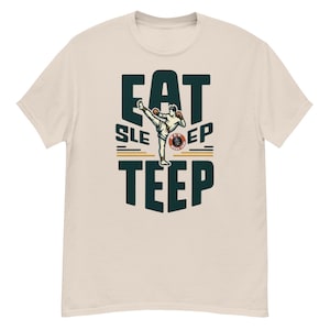 Puede incluir: Una camiseta beige con un gráfico verde que dice "Eat Sleep Teep". El gráfico presenta una caricatura de una persona haciendo una patada teep en una postura de Muay Thai.