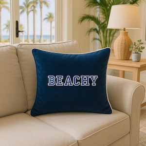 May include: A navy blue square pillow with the word "BEACHY" in white block letters. The pillow has a white border and is placed on a beige sofa. The background includes a window with palm trees and a lamp.