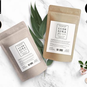 Puede incluir: Dos bolsas de papel marrón con etiquetas blancas. Las etiquetas tienen el texto "LUSH AURA Bath Salts" y enumeran los ingredientes y las instrucciones. Las bolsas están sobre una superficie de mármol blanco.