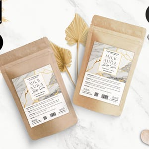 Puede incluir: Dos bolsas de papel marrón con etiquetas blancas. Las etiquetas tienen un diseño de mármol con detalles dorados. El texto en las etiquetas dice "Milk Aura Bath Salts" y "Chamomile & Clay" en una fuente de letra dorada. Las bolsas están sobre una superficie de mármol blanco.