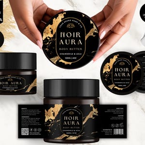 Op de afbeelding: Een zwarte en gouden body butter pot met het label "Noir Aura Body Butter Chamomile & Acai 120ml | 4oz". De pot staat op een wit marmeren oppervlak.