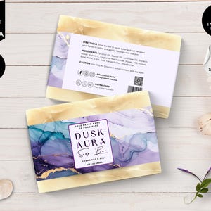 Puede incluir: Una etiqueta de jabón con un diseño abstracto morado y dorado. La etiqueta dice "Dusk Aura Soap Bar" y "Chamomile & Acai" con un peso de 4oz / 113 gramos. La parte posterior de la etiqueta tiene instrucciones, ingredientes y una advertencia.