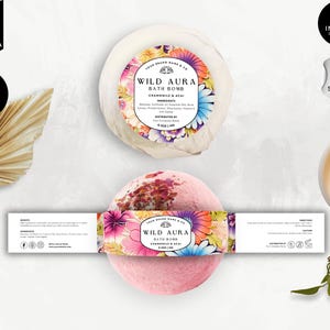 Puede incluir: Dos bombas de baño con etiquetas florales, una blanca y otra rosa, se muestran con una vela encendida y hojas decorativas. Las etiquetas dicen "Wild Aura Bath Bomb" con "Chamomile & Acai" y detalles de ingredientes.