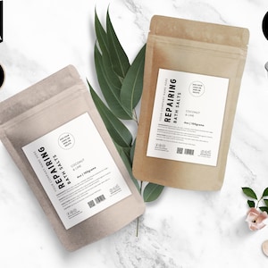 Peut inclure: Deux sachets en papier kraft avec des étiquettes blanches. Les étiquettes portent l'inscription "REPAIRING BATH SALTS" et "COCONUT & LIME". Les sachets sont posés sur une surface en marbre blanc avec des feuilles vertes et une fleur rose.
