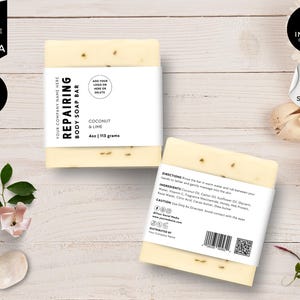 Op de afbeelding: Twee witte stukken zeep met een wit etiket. Het etiket zegt "Repairing Body Soap Bar" en "Coconut & Lime" met het gewicht vermeld als "4oz | 113 grams". Het etiket heeft ook een plek om een logo toe te voegen. De zeep ligt op een lichte houten achtergrond.