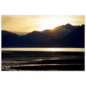 May include: A scenic landscape photograph of a mountain range at sunset. The sun is setting behind the mountains, casting a golden glow over the water and the sky. The foreground shows a dark, textured shoreline.
