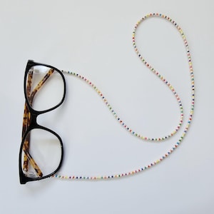 May include: Black-framed spectacles with tortoiseshell arms are attached to a beaded eyeglass chain. The chain features small, colourful beads in a rainbow pattern. The glasses and chain are set against a white backdrop.