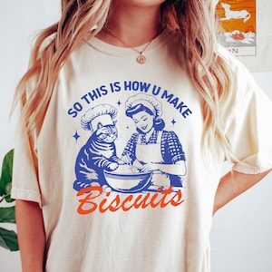 May include: Cream-colored t-shirt with a blue and orange graphic. The graphic features a cat and a woman in chef hats, mixing ingredients in a bowl. Text reads "SO THIS IS HOW U MAKE Biscuits."
