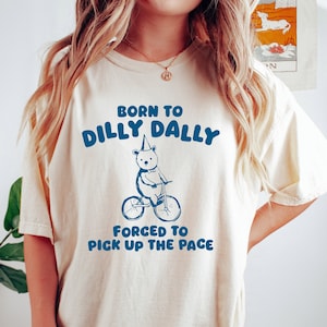 May include: A white t-shirt with a blue graphic of a bear riding a bicycle. The graphic says "Born to Dilly Dally" and "Forced to Pick Up the Pace".