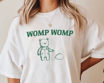 Puede incluir: Camiseta blanca de gran tamaño con un oso de dibujos animados verde que sostiene un globo y el texto "WOMP WOMP" impreso en la parte delantera.