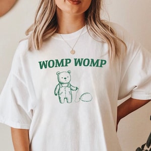 May include: White oversized t-shirt with a green cartoon bear holding a balloon and the text "WOMP WOMP" printed on the front.