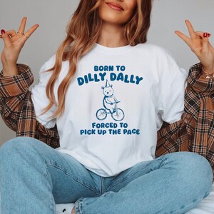 May include: White t-shirt with a blue graphic of a bear wearing a party hat riding a bicycle. The text "Born to Dilly Dally" and "Forced to Pick Up the Pace" is printed on the shirt.