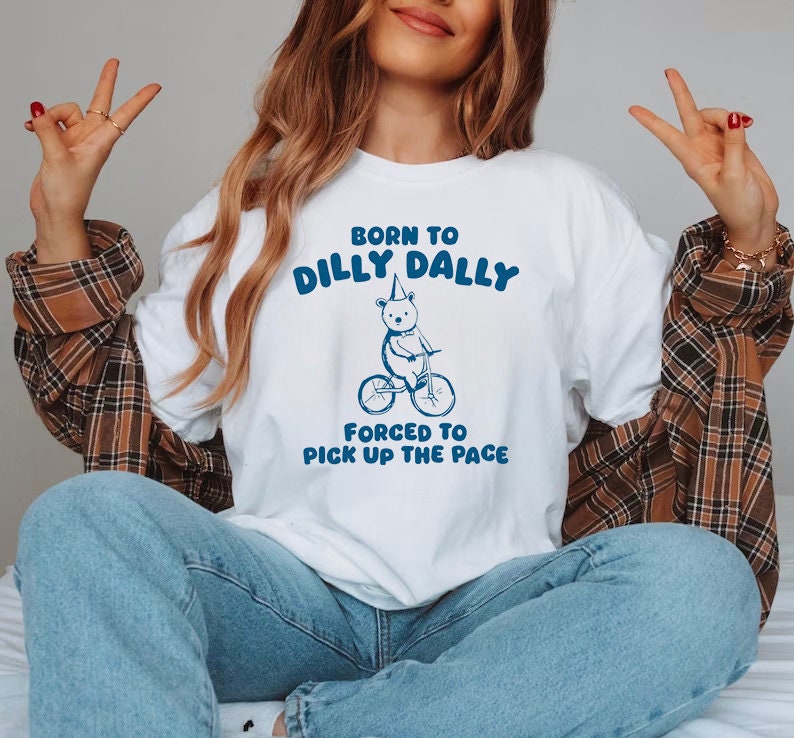 May include: White t-shirt with a blue graphic of a bear wearing a party hat riding a bicycle. The text "Born to Dilly Dally" and "Forced to Pick Up the Pace" is printed on the shirt.