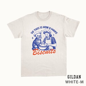May include: White t-shirt with a retro graphic of a woman and a cat wearing chef hats, mixing ingredients in a bowl. The text reads "SO THIS IS HOW U MAKE Biscuits" in blue and orange. The shirt is a Gildan White-M.