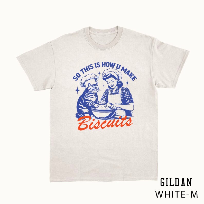 May include: White t-shirt with a retro graphic of a woman and a cat wearing chef hats, mixing ingredients in a bowl. The text reads "SO THIS IS HOW U MAKE Biscuits" in blue and orange. The shirt is a Gildan White-M.
