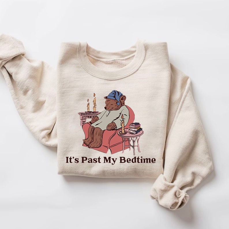 May include: A beige sweatshirt with a graphic of a bear wearing a blue sleep cap and pajamas, sitting in a red armchair with a lit candle. The text "It's Past My Bedtime" is printed below the graphic.