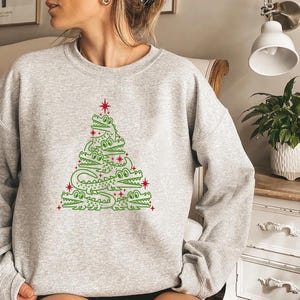 May include: A grey sweatshirt featuring a green alligator Christmas tree design. The tree is made up of alligators stacked on top of each other, with a red star at the top.