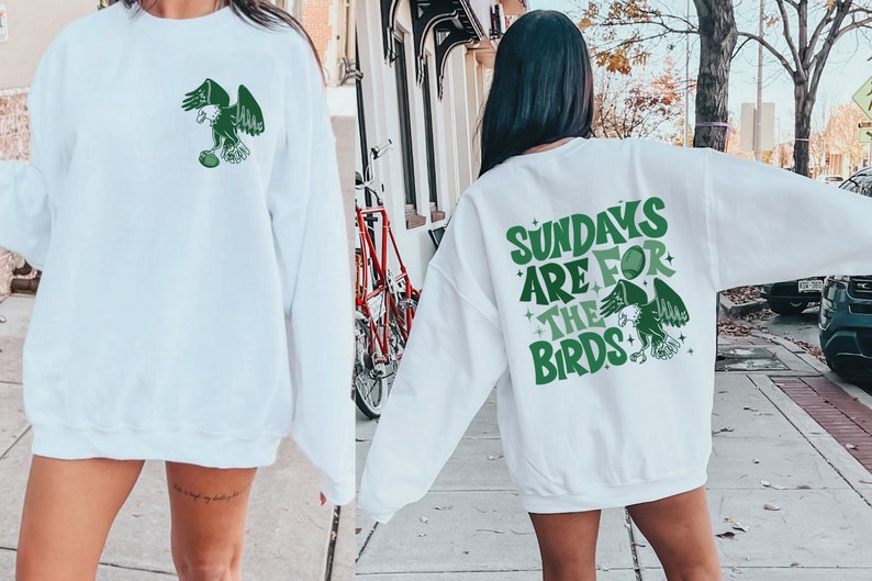Op de afbeelding: Twee witte sweatshirts met groene en witte afbeeldingen. E&eacute;n sweatshirt heeft een afbeelding van een adelaar die een voetbal vasthoudt. Het andere sweatshirt heeft de tekst "Sundays are for the birds" met een afbeelding van een adelaar die een voetbal vasthoudt.