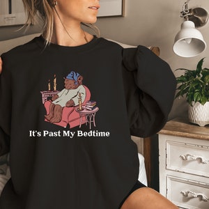 May include: Black sweatshirt with a cartoon illustration of a bear wearing a nightcap and pajamas, sitting in a chair by a fireplace. The text "It's Past My Bedtime" is printed below the illustration.