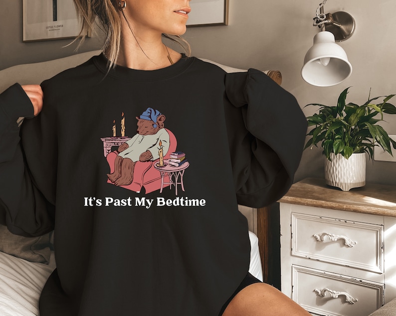 May include: Black sweatshirt with a cartoon illustration of a bear wearing a nightcap and pajamas, sitting in a chair by a fireplace. The text "It's Past My Bedtime" is printed below the illustration.