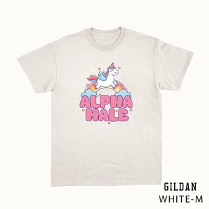 Puede incluir: Una camiseta blanca con un unicornio de dibujos animados volando sobre un arco&iacute;ris y el texto "ALPHA MALE" en letras rosas.