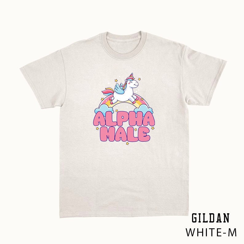 Puede incluir: Una camiseta blanca con un unicornio de dibujos animados volando sobre un arco&iacute;ris y el texto "ALPHA MALE" en letras rosas.