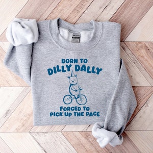 May include: Gray sweatshirt with the text "Born to dilly dally, forced to pick up the pace" in blue. A cartoon bear wearing a party hat rides a bicycle.