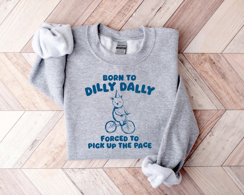 May include: Gray sweatshirt with the text "Born to dilly dally, forced to pick up the pace" in blue. A cartoon bear wearing a party hat rides a bicycle.