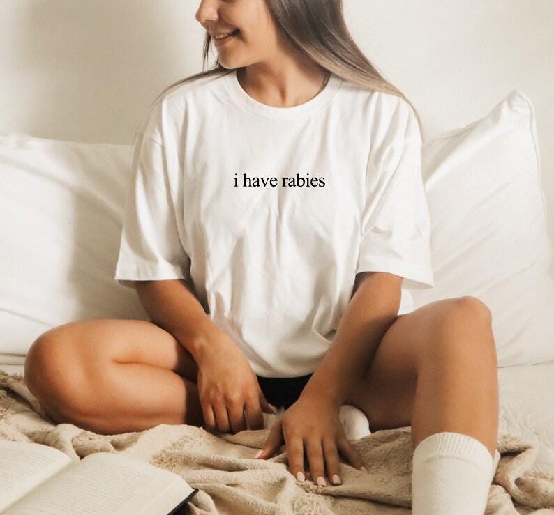 May include: A white t-shirt with the text "i have rabies" printed on the front. The shirt is being worn by a person sitting on a bed.