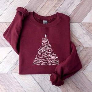 May include: A maroon sweatshirt featuring a white design of an alligator Christmas tree. The tree is made up of alligators stacked on top of each other, with a star on top.