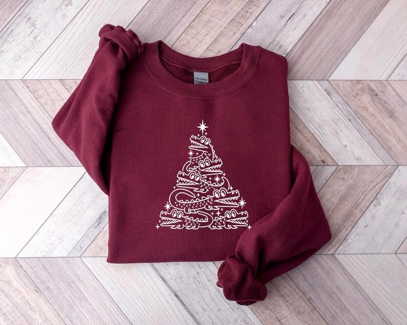 May include: A maroon sweatshirt featuring a white design of an alligator Christmas tree. The tree is made up of alligators stacked on top of each other, with a star on top.