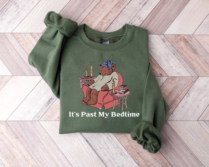 May include: A green sweatshirt with a cartoon bear wearing a nightcap and sitting in a chair with a book and a lit candle. The text "It's Past My Bedtime" is printed below the image.