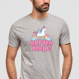 Puede incluir: Una camiseta gris jaspeada con un unicornio de dibujos animados volando sobre un arco&iacute;ris y el texto "ALPHA MALE" en letras rosas.