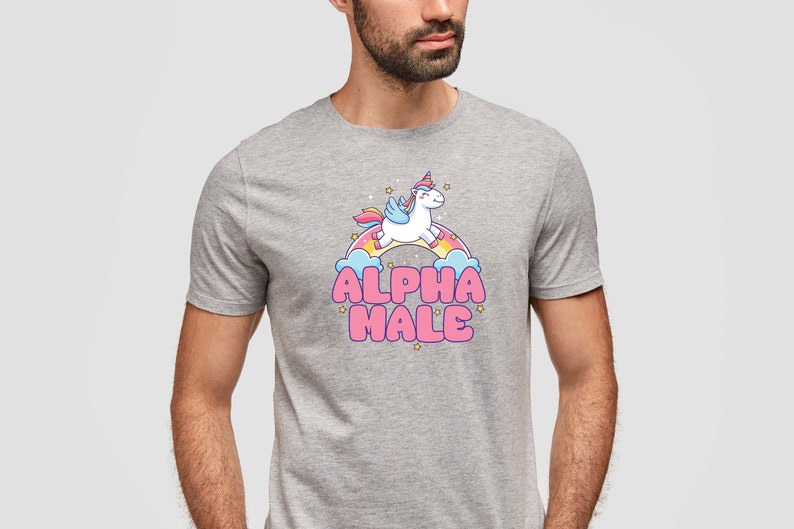 Puede incluir: Una camiseta gris jaspeada con un unicornio de dibujos animados volando sobre un arco&iacute;ris y el texto "ALPHA MALE" en letras rosas.