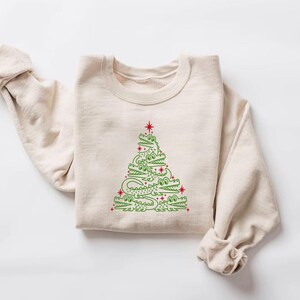 May include: A beige sweatshirt featuring a green alligator Christmas tree design. The tree is made up of alligators stacked on top of each other, with red stars on the branches.