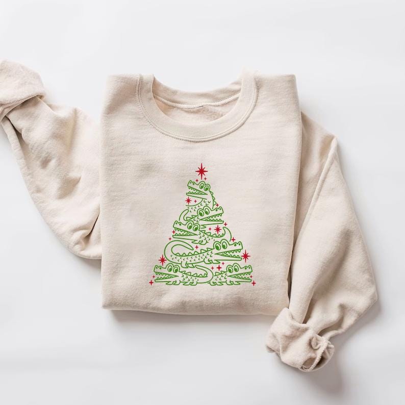 May include: A beige sweatshirt featuring a green alligator Christmas tree design. The tree is made up of alligators stacked on top of each other, with red stars on the branches.